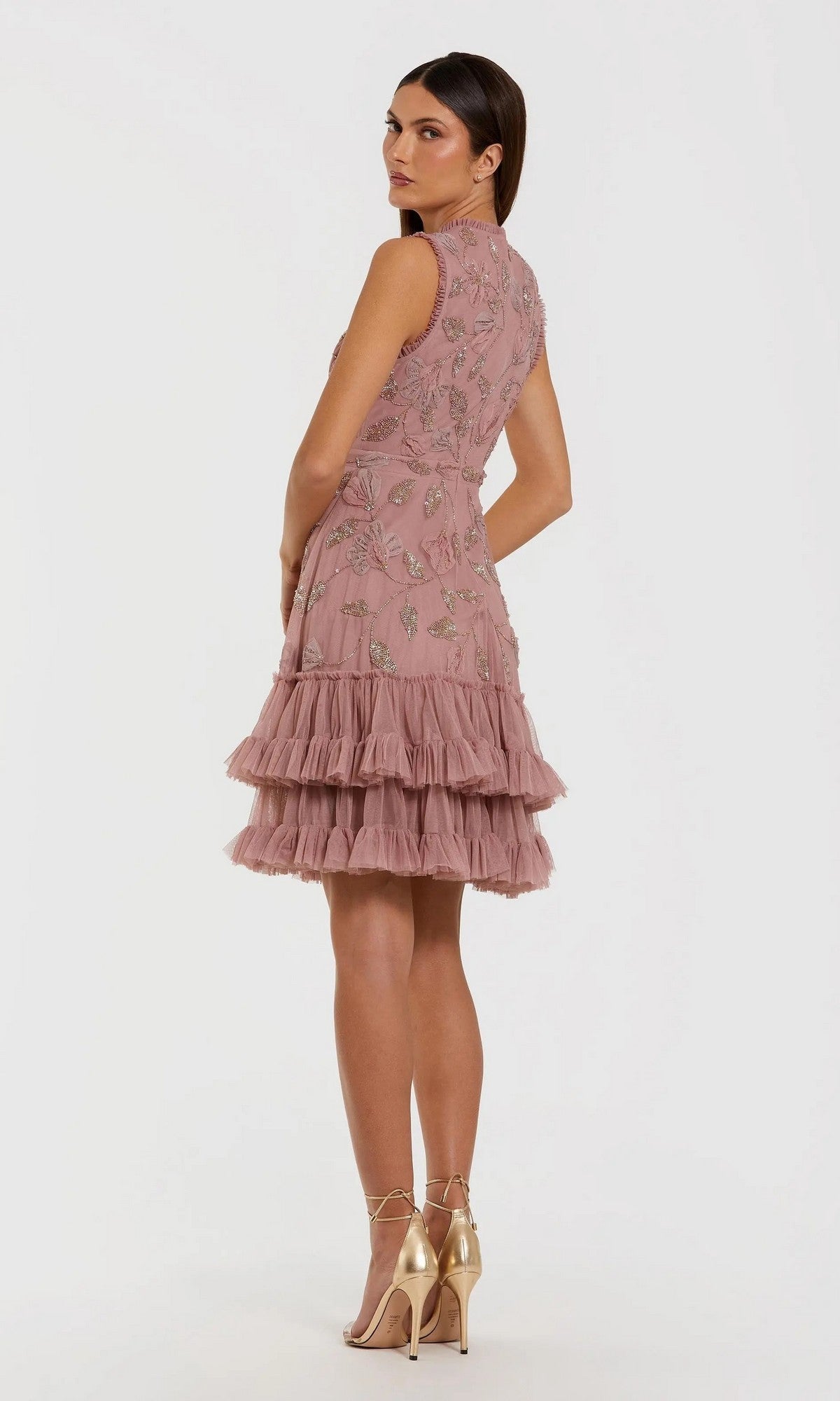 A woman wearing the Mac Duggal 9403 Short Party Dress—a sleeveless, knee-length pink fit and flare design featuring floral embroidery and tiered ruffles—faces slightly away while pairing it with gold high-heeled sandals against a plain white background.